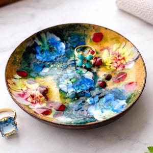 May include: A decorative, round trinket dish with a floral design in shades of blue, yellow, and red. The dish holds a gold ring with a turquoise flower design and a gold ring with a blue gemstone.