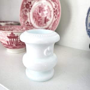 May include: A small, white, decorative vase with a rounded base and a flared top. The vase has a detailed, beaded rim and a small, curved design on the side. The background includes pink and white patterned plates and bowls.