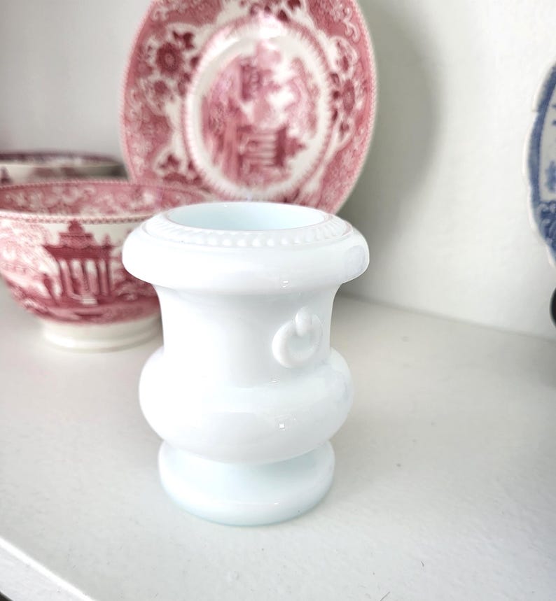 May include: A small, white, decorative vase with a rounded base and a flared top. The vase has a detailed, beaded rim and a small, curved design on the side. The background includes pink and white patterned plates and bowls.