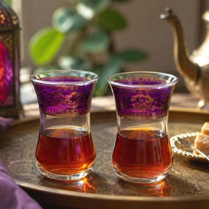 May include: Two clear glass tea cups with a tapered waist, each featuring a wide band of deep purple glass at the rim decorated with intricate gold filigree patterns. Both glasses are filled with amber-colored tea and sit on a round metal tray.