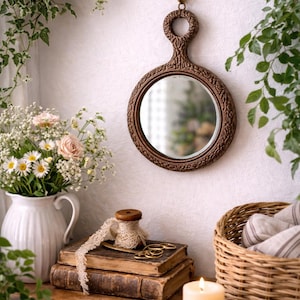 May include: A decorative, round brown mirror with an ornate handle hangs on a white wall. A white pitcher with flowers, old books, a basket, and a lit candle are on a wooden surface. Green plants frame the scene.