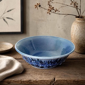 May include: A blue ceramic bowl with a textured rim sits on a wooden surface. The bowl has a smooth interior and a darker blue, patterned exterior. A beige vase with dried branches is in the background.