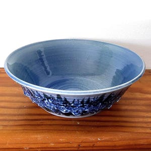 May include: A blue ceramic bowl with a textured, dark blue floral design around the rim. The interior of the bowl is a lighter shade of blue. The bowl is sitting on a wooden surface.