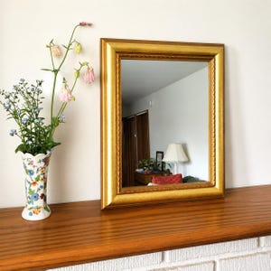 May include: A rectangular gold-framed mirror with a decorative rope-like border. The mirror is displayed on a wooden surface, next to a vase filled with flowers. The vase has a floral pattern.