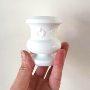 May include: A white ceramic candle holder with a decorative design. The candle holder has a bulbous shape with a detailed top and a small, round base. The holder is held in a hand against a plain background.