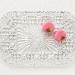 May include: A clear, rectangular glass serving dish with a detailed, textured pattern. Two pink, round candies with green accents rest on the dish. The dish has a decorative, scalloped edge and a starburst design in the center.