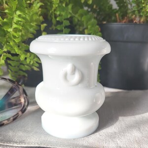 May include: A white ceramic vase with a rounded base and a flared top. The vase features a power button design and a decorative beaded rim. The background includes green plants and a pair of eyeglasses.