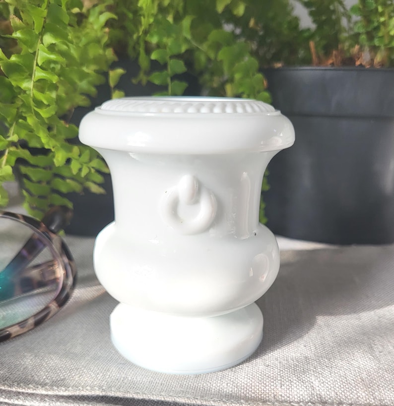 May include: A white ceramic vase with a rounded base and a flared top. The vase features a power button design and a decorative beaded rim. The background includes green plants and a pair of eyeglasses.
