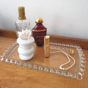 May include: A clear glass tray with a beaded edge holds a collection of items. A perfume bottle with a gold cap, a red perfume bottle with a gold cap, a white cotton swab holder, a gold lipstick tube, and a pearl necklace are visible.