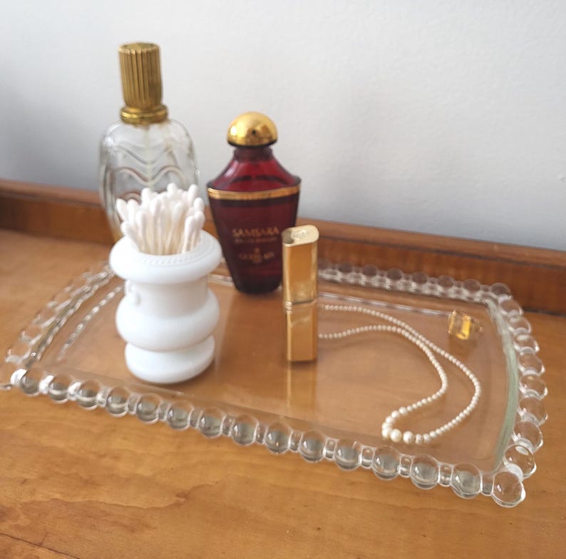 May include: A clear glass tray with a beaded edge holds a collection of items. A perfume bottle with a gold cap, a red perfume bottle with a gold cap, a white cotton swab holder, a gold lipstick tube, and a pearl necklace are visible.