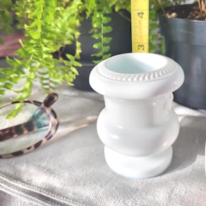 May include: A white, decorative vase with a bulbous shape and a textured rim. The vase is approximately 3 inches tall. The background includes green plants and a measuring tape.