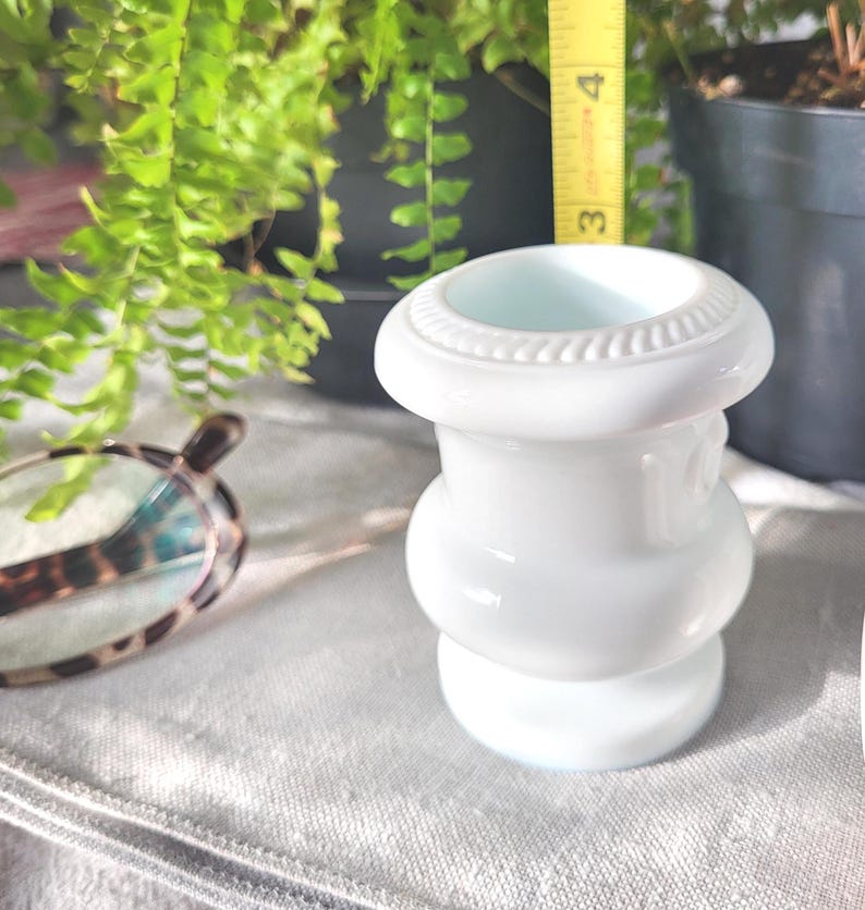 May include: A white, decorative vase with a bulbous shape and a textured rim. The vase is approximately 3 inches tall. The background includes green plants and a measuring tape.