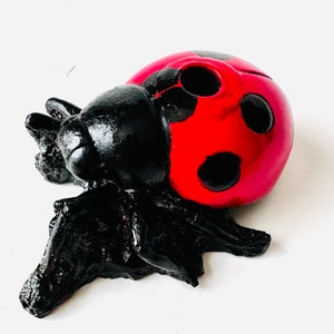 May include: A decorative ladybug figurine. The ladybug has a red shell with black spots and a black head and body. The figurine is made of a glossy material and sits on a white surface.