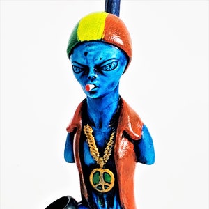 May include: A blue ceramic pipe shaped like an alien wearing a red, yellow, and green hat, a red jacket, and a gold chain with a peace sign pendant.