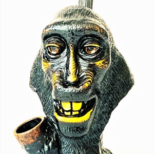 May include: A black ceramic pipe shaped like a monkey's head with a yellow painted mouth and eyes. The pipe has a brown bowl.