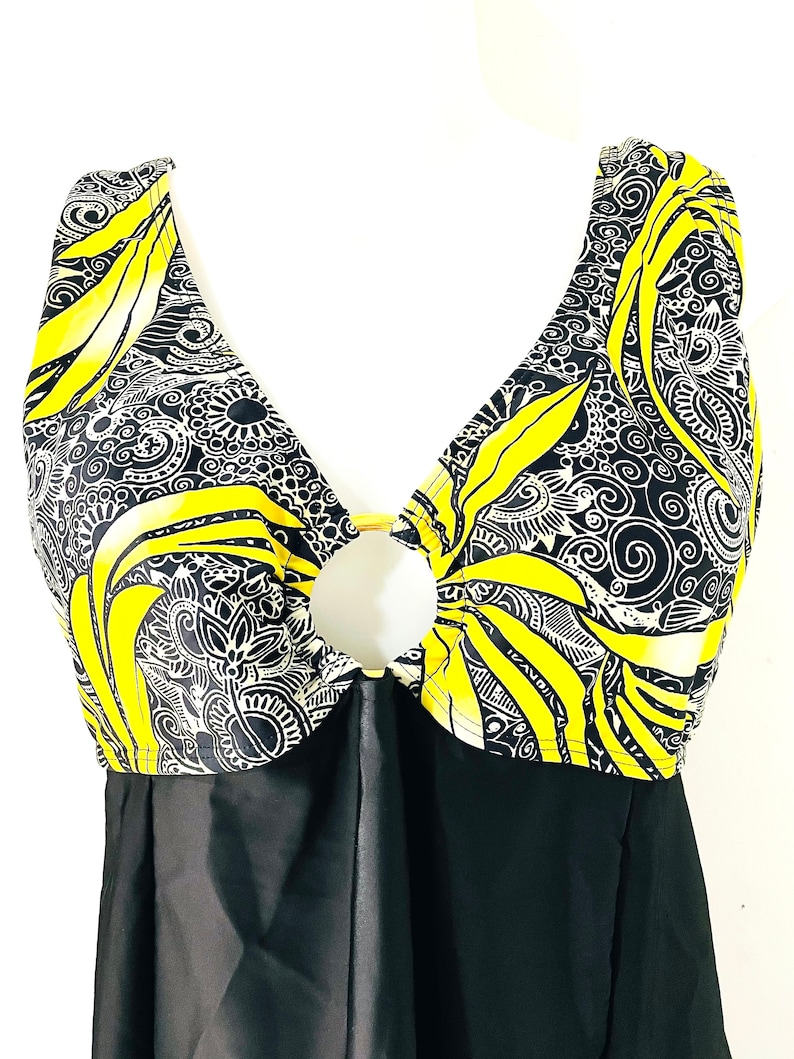 Puede incluir: Un ba&ntilde;ador de una pieza con falda negra y top con dise&ntilde;o estampado en amarillo, negro y blanco. El top tiene un escote pronunciado con un detalle de anillo dorado. El ba&ntilde;ador se exhibe en un maniqu&iacute; blanco.