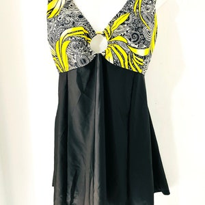 Puede incluir: Un traje de ba&ntilde;o negro con falda. La parte superior presenta un dise&ntilde;o de cuello en V con un detalle circular y una tela estampada amarilla y negra. El patr&oacute;n incluye remolinos y formas de hojas.