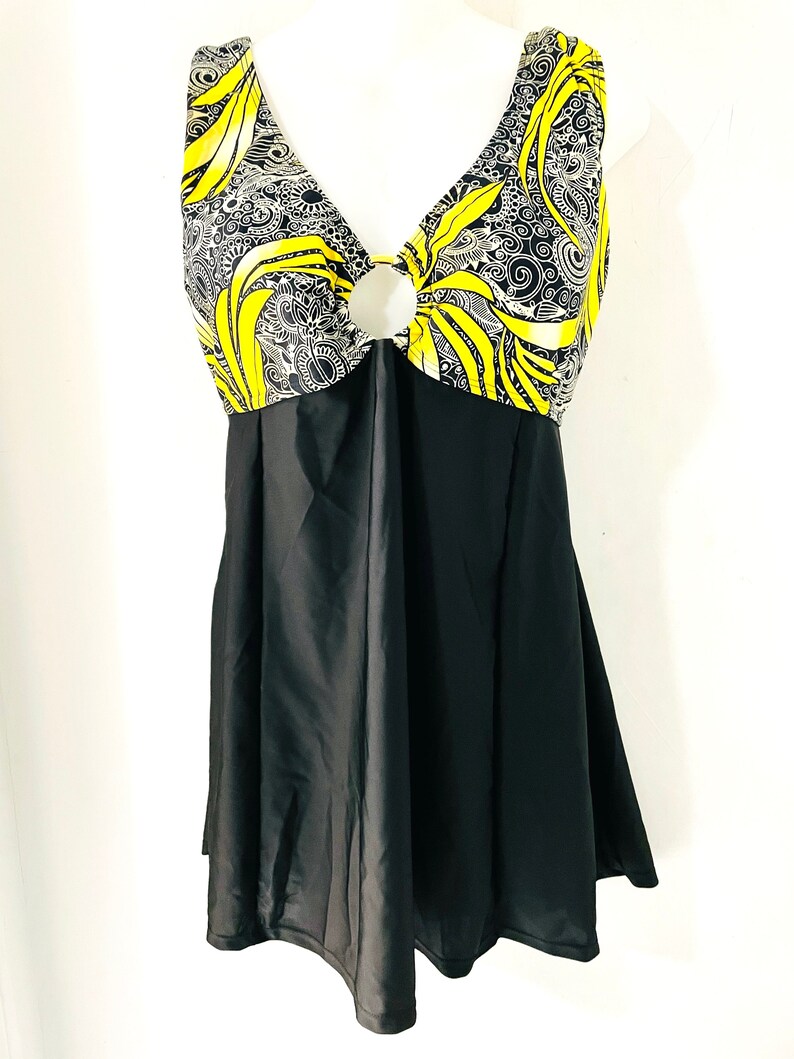 Puede incluir: Un traje de ba&ntilde;o negro con falda. La parte superior presenta un dise&ntilde;o de cuello en V con un detalle circular y una tela estampada amarilla y negra. El patr&oacute;n incluye remolinos y formas de hojas.