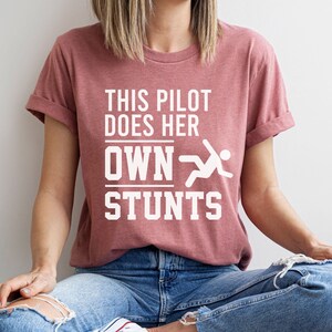 May include: A dusty rose t-shirt with the text "THIS PILOT DOES HER OWN STUNTS" in white block letters. A white graphic of a person in motion is next to the text. The shirt is worn with blue jeans.