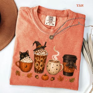 Può includere: Una t-shirt girocollo color corallo con una stampa grafica raffigurante un gatto nero in una tazza, un caffè freddo con un cappello da strega, una tazza fumante con stelle e un bicchiere da caffè con un pipistrello. Etichetta Comfort Colors e testo YAM.