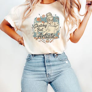 以下が含まれることがあります： クリーム色のTシャツで、メガネをかけてお団子ヘアの人物が本に絵を描いているグラフィックイラストが描かれています。テキストは「Sketchy But Artistic」とあり、筆、絵の具のチューブ、インク瓶などの画材が落ち着いた色調で周囲に配置されています。
