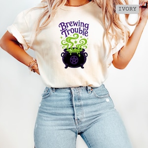 May include: An ivory short-sleeve t-shirt featuring a graphic of a black cauldron with a purple pentagram, green bubbling liquid, and the text Brewing Trouble in purple script. The shirt is paired with light blue denim jeans.