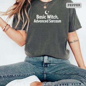 Può includere: Una t-shirt a maniche corte color grigio antracite con testo bianco che recita Basic Witch, Advanced Sarcasm e un'icona a forma di mezzaluna e stella sopra le parole. La maglia presenta un girocollo e una vestibilità ampia, abbinata a jeans blu.