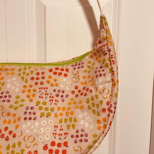 Puede incluir: Un bolso de hombro en forma de media luna de color rosa claro con un colorido estampado de frutas. El bolso presenta un diseño acolchado y una cremallera verde. Las ilustraciones de frutas incluyen fresas, manzanas, plátanos y naranjas.