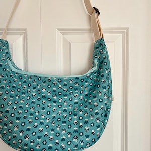 May include: A teal crescent-shaped shoulder bag with a cream-coloured adjustable strap. The bag features a quilted pattern of white and dark teal mushrooms and flowers. The zip is light teal.