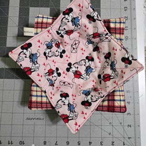 May include: Two square bowl cozies. The top cozy is light pink with a pattern of Mickey and Minnie Mouse, red hearts, and envelopes. The bottom cozy has a plaid pattern in red, cream, and blue. The cozies are on a gray cutting mat.
