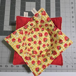 May include: A quilted red square pot holder with a yellow and white checkered strawberry-patterned pot holder on top. The strawberry pot holder is diamond-shaped. The image includes a tag that says "Klima Creations".