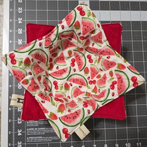 May include: A square fabric bowl cozy featuring a white background with a repeating pattern of red watermelon slices, strawberries, and cherries. The interior is lined with solid red fabric and includes two small beige fabric tags on the sides.