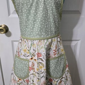 May include: A floral apron with a sage green polka dot bodice and pockets. The apron features a white background with colorful flowers and green trim. The apron is displayed on a mannequin.