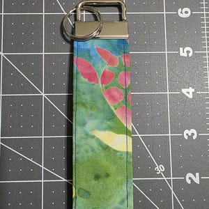 May include: A fabric keychain wristlet featuring a colorful turtle pattern with shades of green, pink, and yellow. The strap is attached to a silver-tone metal hardware clasp and a circular split ring, measuring approximately 5.5 inches in length.