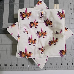 May include: Two white square pot holders with a repeating pattern of unicorn faces. The unicorns have purple horns, orange and purple floral manes, and are set against a white background. The pot holders are layered on top of each other.
