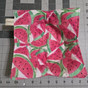 May include: A square, quilted pot holder featuring a watermelon slice pattern in shades of pink, green, and white. The pot holder has a white border and a small tag that reads "Creations".