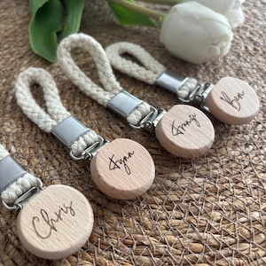 Könnte beinhalten: Vier Schnullerketten mit geflochtenen cremefarbenen Baumwollschlaufen, silberfarbenen Metallclips und runden hellen Holzscheiben. Jede Holzscheibe ist mit einem Namen in Schreibschrift graviert: Chris, Fynn, Franzi und Ben.