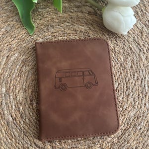 Könnte beinhalten: Brauner Leder-Reisepasshalter mit einer genähten Bordüre. Auf der Vorderseite befindet sich eine schwarze Strichzeichnung eines Oldtimer-Campingbusses. Er liegt auf einer gewebten, beigefarbenen Oberfläche, mit weißen Blumen und grünen Blättern im Hintergrund.