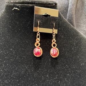 May include: A pair of handcrafted earrings with a copper wire design. Each earring features a red and white speckled stone, suspended from a series of copper wire loops. The earrings are displayed on a black jewelry stand.
