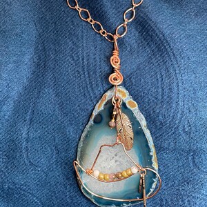 May include: A copper chain necklace with a pendant made from a slice of agate. The agate is blue and white with a brown band around the edge. The pendant is wrapped in copper wire and has a small copper feather charm and a few small beads.