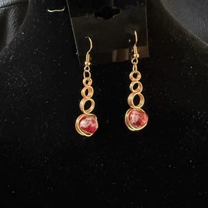 May include: A pair of gold-colored wire earrings. Each earring features a series of connected circles leading to a red, rounded stone at the bottom. The earrings are displayed against a black background.