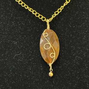 May include: A gold-colored necklace with a pendant featuring a brown, oval-shaped stone. The stone is wire-wrapped with gold-colored wire in a decorative pattern. A small gold bead hangs below the stone, and the chain is a delicate link style.