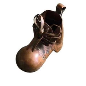 May include: A small, copper-colored boot-shaped container. The boot has a rounded toe, laces, and a loop at the top. The container is empty and appears to be made of metal. The boot is a decorative item.