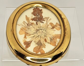 May include: A small, round, gold-toned picture frame with a dried flower arrangement. The arrangement features various dried flowers and leaves in shades of brown and beige, set against a cream-colored background. The frame has a slightly worn appearance.