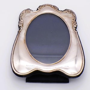 May include: A silver-toned picture frame with an oval centre opening and a decorative, scalloped edge featuring embossed floral details. The frame has a smooth, reflective metallic surface and is mounted on a dark, velvet-like backing material.