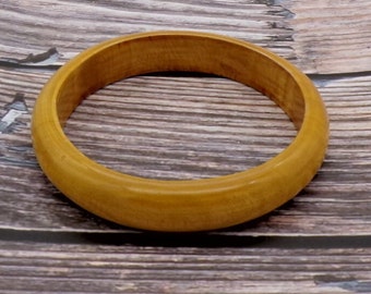 May include: A light brown wooden bangle bracelet. The bracelet is circular and has a smooth, polished finish. The wood grain is visible, adding texture to the piece. The bracelet is set against a wooden background.
