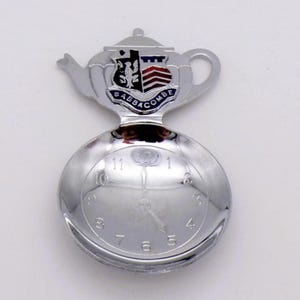 Puede incluir: Una cuchara de recuerdo de metal plateado con un mango en forma de tetera que presenta un escudo colorido y el texto BABBACOMBE. El cuenco de la cuchara es circular y está grabado con una esfera de reloj con números del uno al doce.