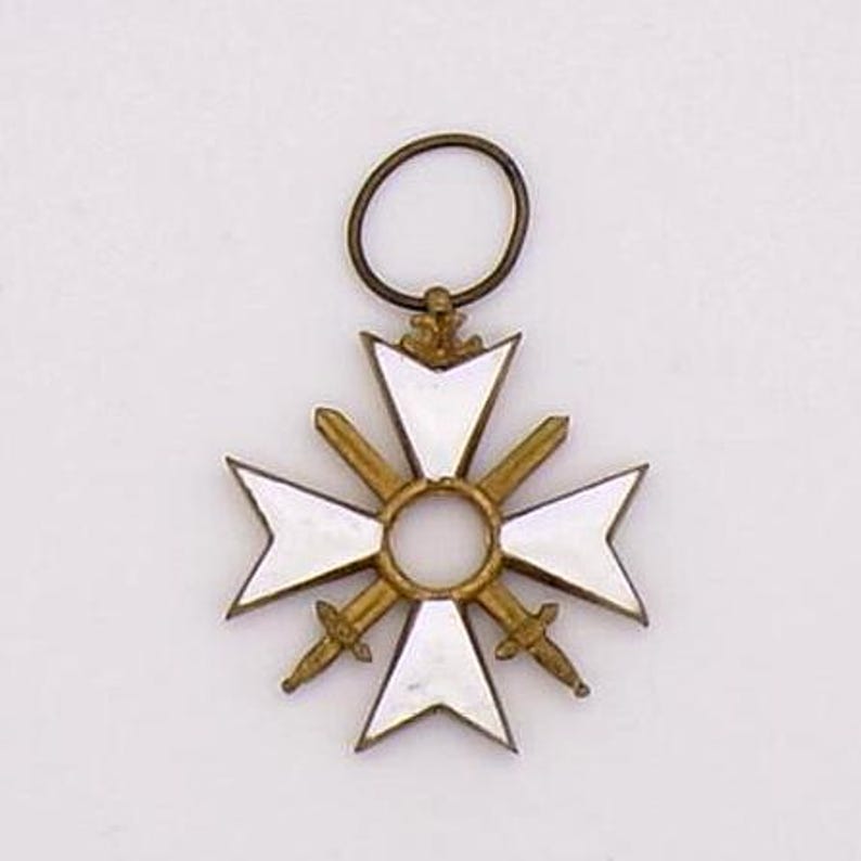 May include: A gold-toned pendant in the shape of a cross with white enamel sections and two swords. The pendant has a circular loop at the top for hanging. The cross design is centered with a circular element.