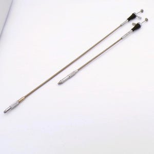 May include: Two silver and bronze camera cable releases. The cables are thin and flexible, with a metal tip on one end and a black rubber button on the other. The background is white.