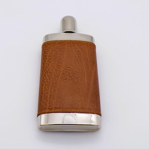May include: A rectangular metal flask featuring a polished silver-tone finish at the top and bottom, with a textured tan leather wrap covering the centre. The leather section displays a small, embossed circular logo in the middle of the front.